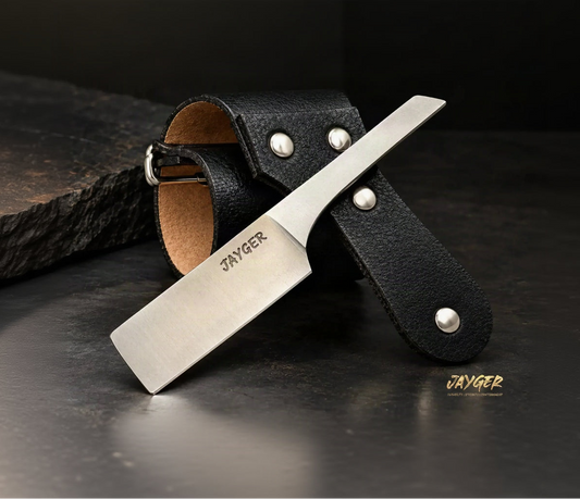 A Jayger kamisori-style knife with a satin-finished silver blade, displayed diagonally over a black leather strap on a dark wooden surface. Above the knife, four gold icons highlight key features: carbon steel, kamisori style, shave ready, and leather strap included.